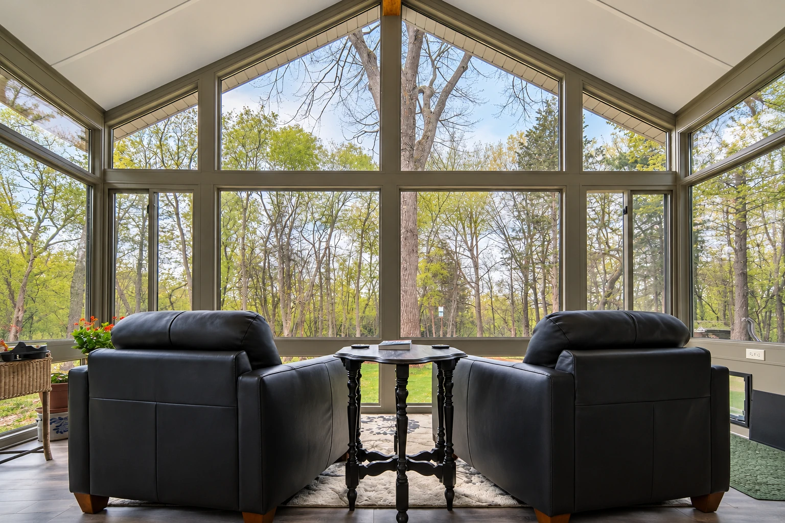Four-season sunroom with soaring grid-framed windows, two black armchairs, and a wooded view.