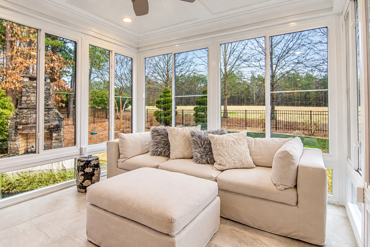 Bright all-season sunroom with wraparound windows, a cream sofa and ottoman, and a view of the yard.