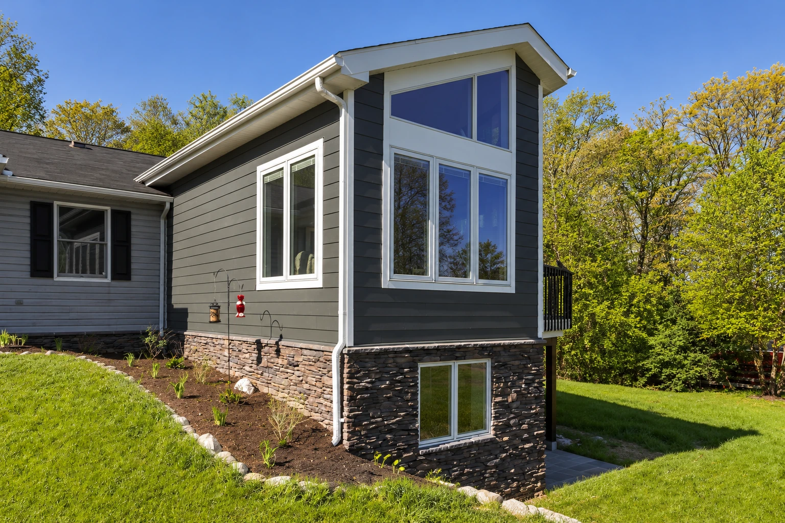 Custom sunroom addition with dark siding, a tall bank of windows and a stone-clad foundation beside a wooded yard.