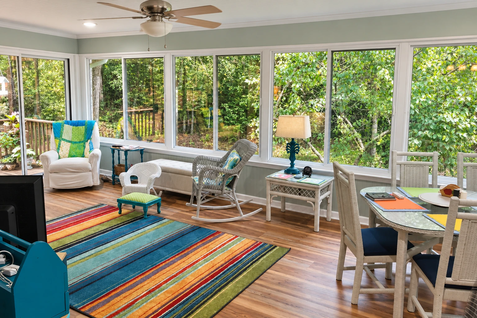 A spacious sunroom has white-framed windows overlooking trees, with wicker chairs, a dining table and a colorful striped rug.