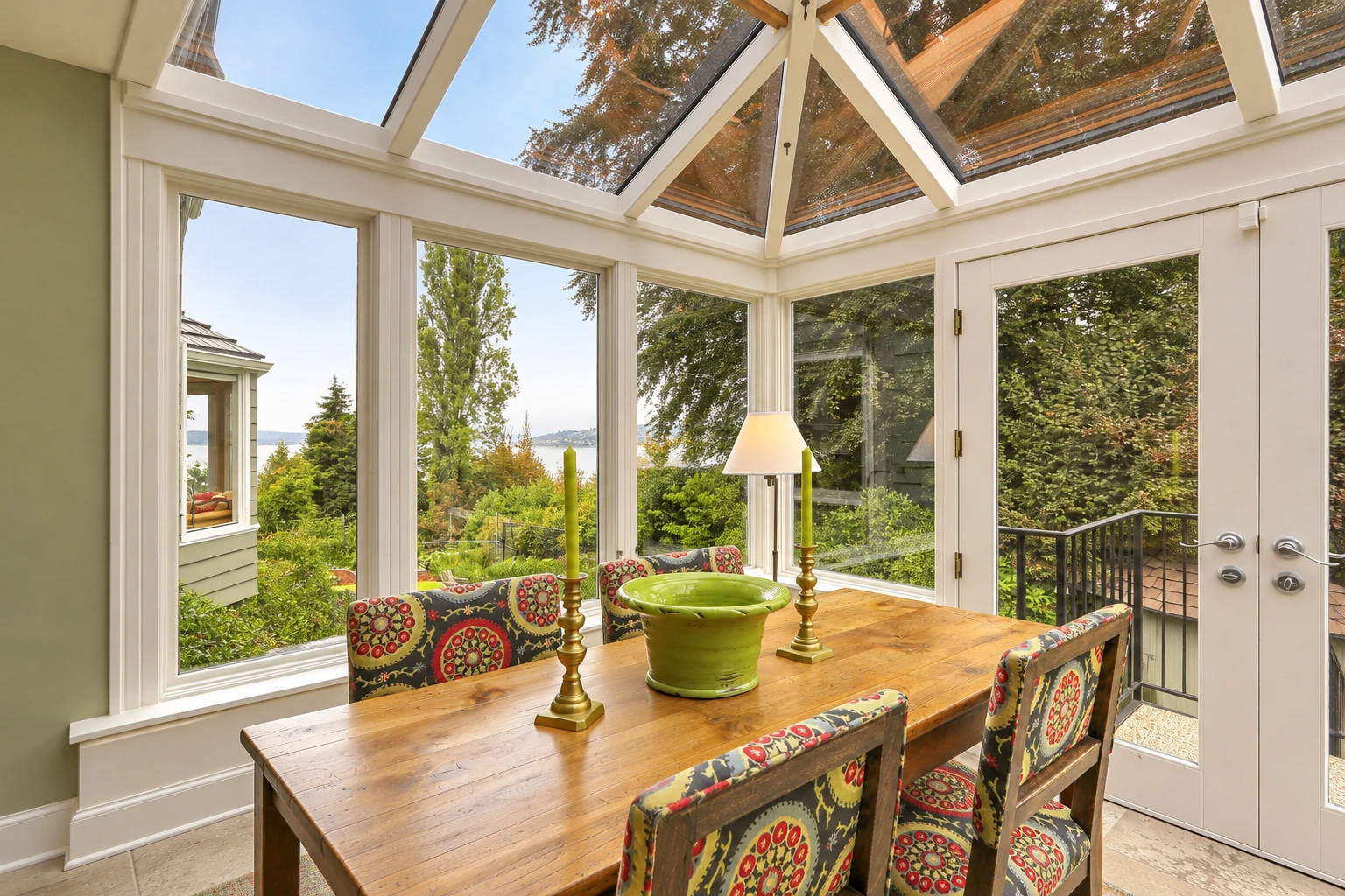 A sunroom dining area with a glass-paneled roof, wooden table, patterned chairs and views of a leafy yard.