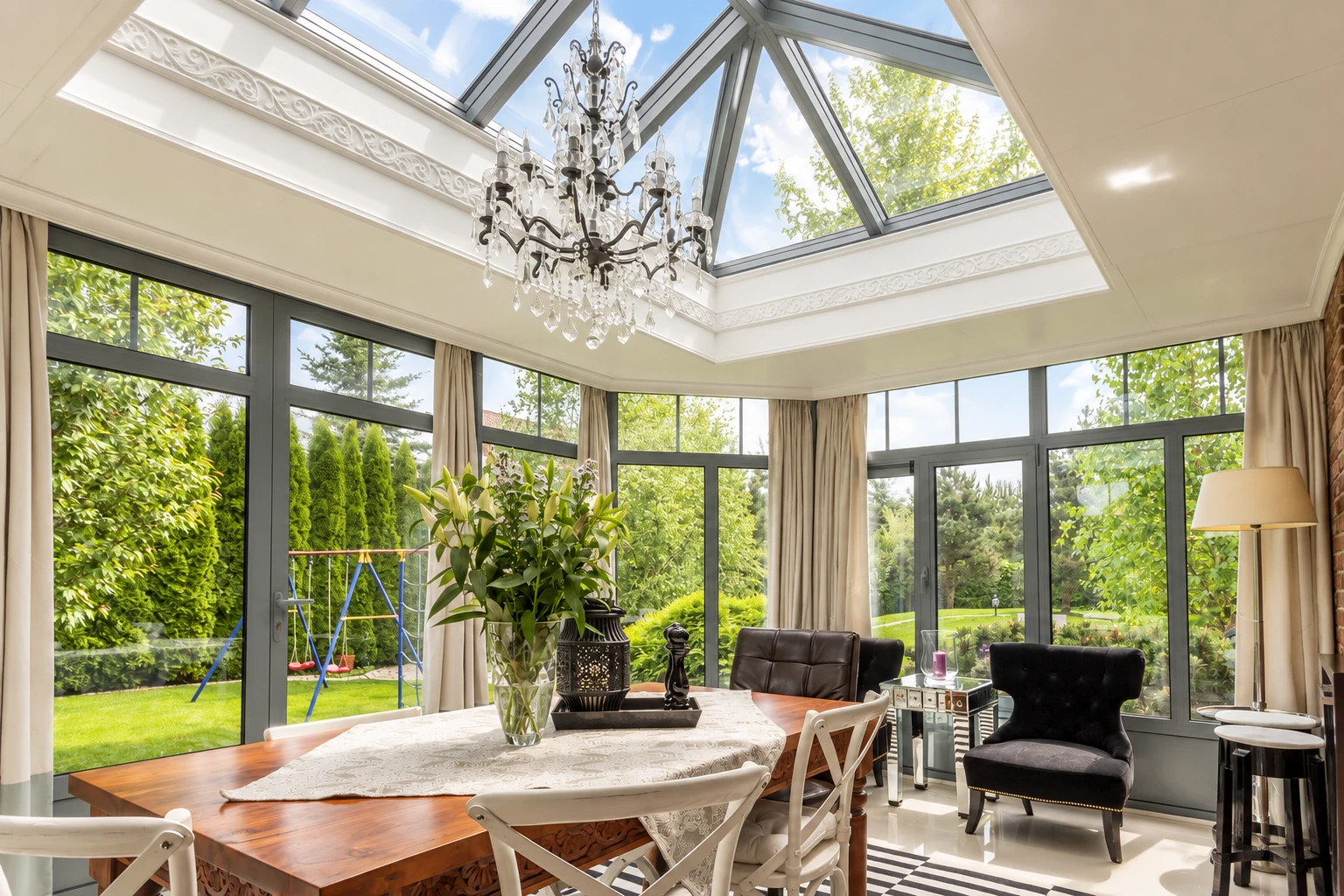 A bright sunroom with a peaked glass ceiling, dark-framed windows, chandelier and dining furniture overlooking a garden.